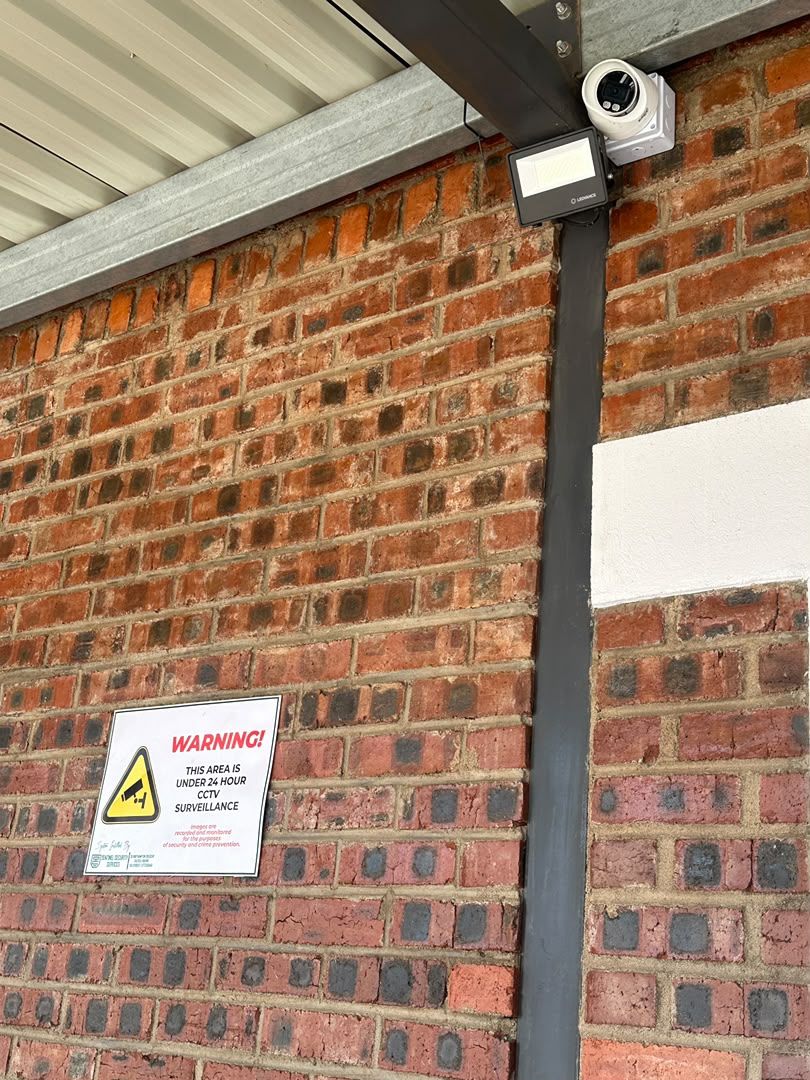 A dome camera and floodlight under an eave, with the warning sign below